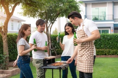 asian man cooking barbeque grill and sausage for a group of friends to eat party in garden at home. group of friends having outdoor garden barbecue laughing with alcoholic beer drinks