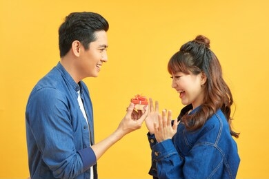 love, couple, proposal and people concept - man giving diamond engagement ring in little red box to happy woman over  yellow background