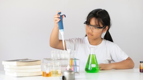 young adorable girl holding a chemistry glassware while doing a scientific experiment and sitting at the modern white table with white laboratory wall as background.