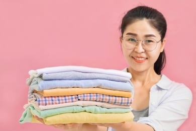 young beautiful asian woman holding stacks of folded washed and dried clothes pile on pink wall background,selective focus