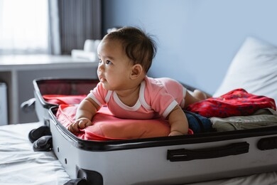 asian baby smiles on his stomach in an open suitcase filled with clothes