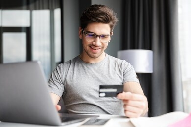 handsome smiling young bearded man wearing casual clothes using laptop computer while sitting at the table at home, showing plastic credit card, shopping