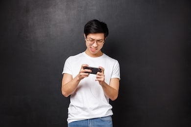 technology, gadgets and people concept. portrait of concentrated, focused asian guy trying kill mob in mobile game, passing arcade or street races, holding smartphone horizontally and grimacing