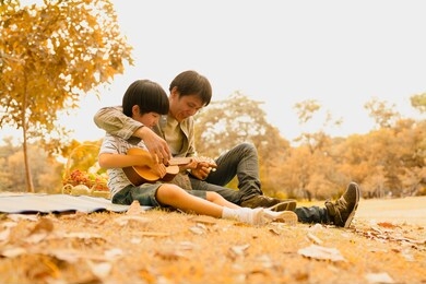 asian father teaching son to play ukulele, spending time together at park in autumn afternoon sunlight, the concept of family outdoor, daddy hero and son, happy time together, father's day, parenting.