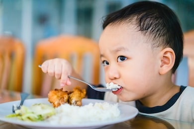 cute little asian kid boy hungry eating rice with fried chicken in table dinner in kitchen at home.child boy holding spoon and eat food with himself.child development, happy meal, hungry face concept.