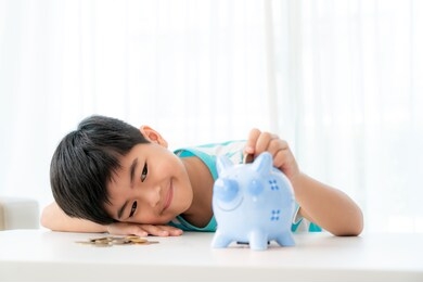 little asian boy insert a coin into blue piggy bank in white table at living room at home for the kid saving, investment money for his business future concept