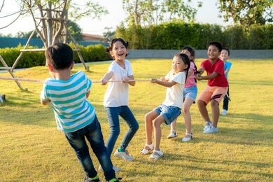 group of happy young asian children playing tug of war or pull rope together outside in city park playground in summer day. children and recreation concept. multi-ethnic children group, outdoor.
