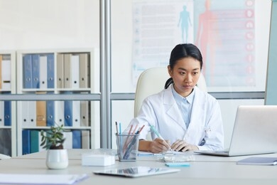 portrait of female asian doctor using laptop while sitting at workplace in medical office interior, copy space