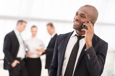 businessman on the phone. cheerful young african man in formalwear talking on the mobile phone and smiling while his colleagues standing on background