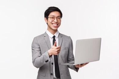 waist-up portrait of satisfied, happy and pleased young asian male entrepreneur, it programmer in suit, introduce his new project, show thumbs-up in approval and holding laptop, white background