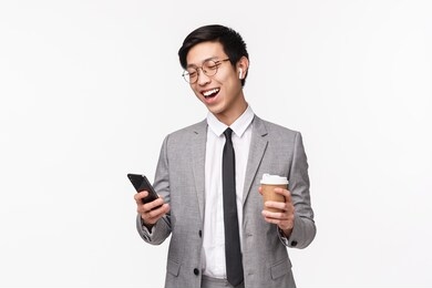 office workers, business and lifestyle concept. waist-up portrait of lucky cheerful asian male entrepreneur calling friend using wireless earphones, hold smartphone and take-away coffee, smiling