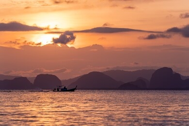 the silhouette of a faraway thai long tail fishing boat sailing past distant mountains during a golden sunset near krabi, thailand