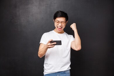 technology, gadgets and people concept. happy joyful lucky asian guy fist pump in celebration, hold mobile phone and smiling say yes, triumphing passed game or level, black background