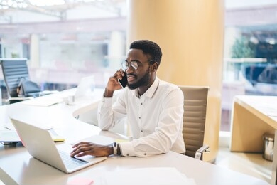 dark skinned man in optical eyeglasses working on startup project research business information on laptop website, black male receiving cellphone call conversation via application for talking