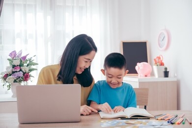 asian cute boy doing his school homework and his mother use laptop at home, he is writing on a book.education technology concept