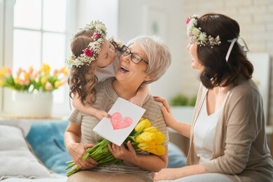 happy mother's day! child daughter is congratulating mom and granny giving them flowers and gift. grandma, mum and girl smiling and hugging. family holiday and togetherness.                         
