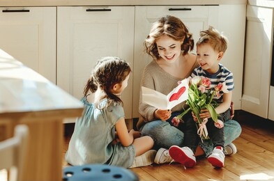 smiling young woman reading greeting card while sitting on floor with cheerful little siblings with bouquet of flowers during mother day
