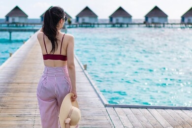 young girl holding hat walking surrounded by beautiful turquoise ocean water. happy traveller in tropical beach vacation. back of young asian woman walking over a wooden jetty in morning sunrise.