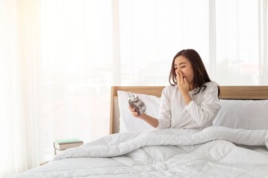 woman in bed trying to wake up with alarm clock. young girl in white on bed. portrait of beautiful young asian girl with alarm clock in hand feeling frustrated on bed after getting up in morning.