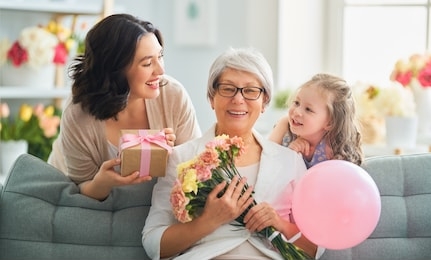 happy mother's day! child daughter is congratulating mom and granny giving them flowers and gift. grandma, mum and girl smiling and hugging. family holiday and togetherness.