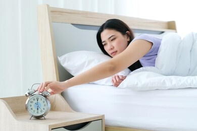 sleeping woman wake up in early morning time (at 6 am) and her hand touching off alarm clock in bedroom. feeling sleepy and not fresh. she sleep on white bed. wake up with alarm clock snooze concept
