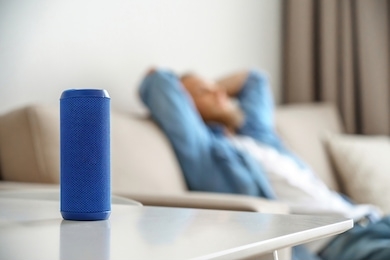small portable wireless speaker assistant on table with man lounge on sofa listening to music. young relaxed guy enjoys mini bluetooth stereo gadget for sound digital assistance at smart home concept.