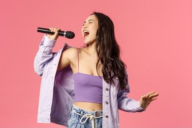 portrait of excited good-looking, carefree asian girl singing with all her heart, put effort to reach that highest note, sing karaoke, close eyes and enjoying performing, standing pink background