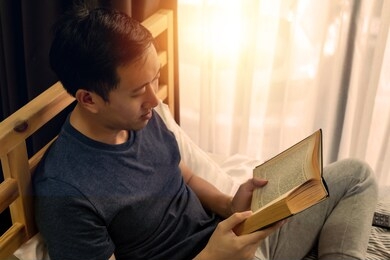 portrait of serene 30s asian man in casual clothing spend free time reading a book inside at home on bed. recreation and leisure time concept