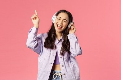 portrait of attractive carefree asian girl chilling and vibing, listening music in headphones, raising finger up while trying sing along and reach highest note in song, pink background
