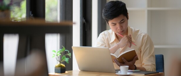 cropped shot of young college student concentrating on book and laptop to prepare his coming exam 