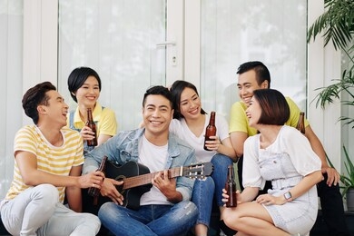 cheerful handsome vietnamese young man playing guitar for his friends drinking and talking at house party