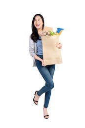 full body of smiling asian woman holding paper shopping bag full of groceries and looking at  empty space aside isolated on white background