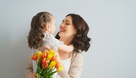 happy mother's day! child is congratulating mom and giving her yellow flowers tulips. mum and girl smiling on light grey background. family holiday and togetherness.                                   