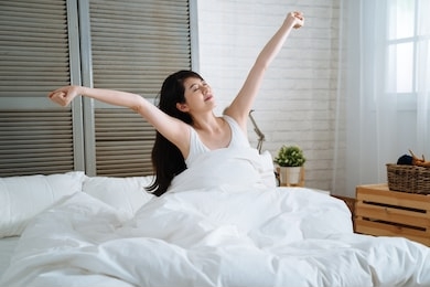 attractive woman stretching in the morning and smiling while get up from bed. young beautiful girl raising arms after wake up in bright cozy bedroom. lady cover by warm white blanket at home