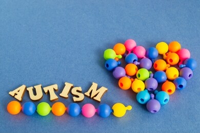 the word autism from simple wooden letters and a puzzle of multi-colored beads is folded in the shape of a heart on a blue background. autism day. autism spectrum disorder concept. space for text
