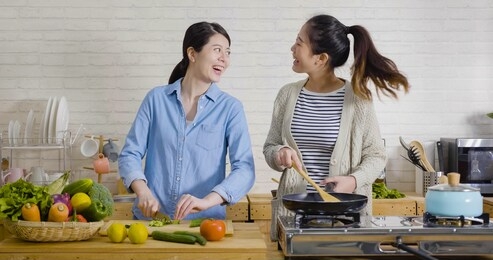 young female friends in kitchen cooking together vegetarian meal. preparing fruit salad healthy food in morning kitchen. happy asian women singing dancing and having fun while cutting and frying