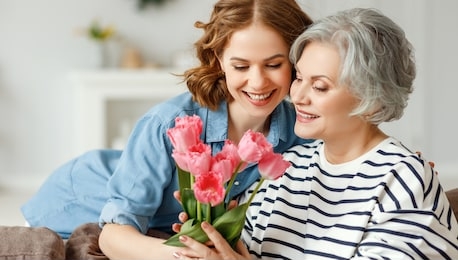 cheerful young female hugging happy aged mother and giving bouquet of tulips while congratulating on mother day at home
