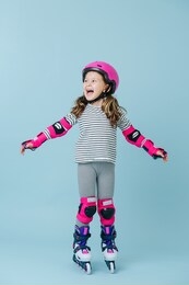 thrilled happy little girl in striped clothes and roller skates with pink protective gear over blue background. her arms wide apart for balance.