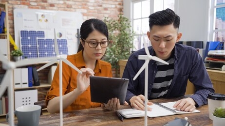 start up small business team in eco friendly office working showing tablet. talented engineers consider wonderful model of powerful windmill generators. asian girl and boy colleagues brainstorming.