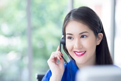 call centre. smiling friendly beautiful face asian woman blue shirt operator working with headset look sideway front of computer screen. technical support customer service agent in an startup office.