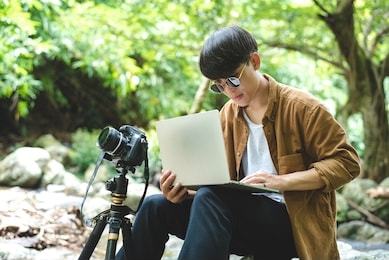 asian man traveler photographer are taking photo and use laptop at beautiful forest. travel photography, blogger, vlogger, camping, work aboard, adventure lifestyle, hipster, tourism concept.