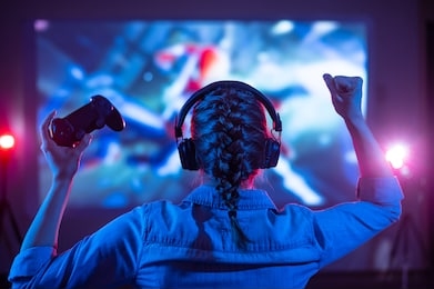 girl in headphones plays a video game on the big tv screen. gamer with a joystick. online gaming with friends, win, prize. fun entertainment. teens play adventure games. back view. neon lighting
