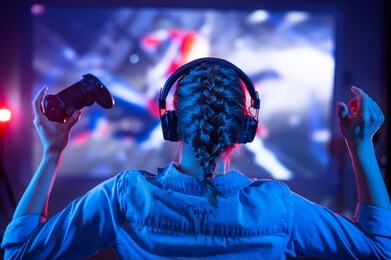young girl in headphones plays a video game on the tv in the dark room. gamer with a joystick. online gaming with friends, competitions, win. fun entertainment. teenagers play puzzles games. back view