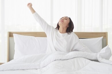 young happy, beautiful asian woman stretching arm, twisting to relax muscles and sitting in white bed while wake up in the morning fresh after get good sleep in bedroom. relaxing and leisure concept.