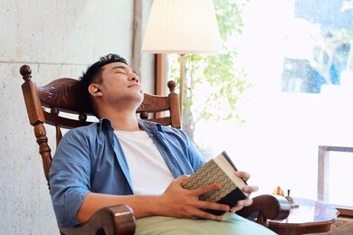 asian man in blue shirt take a nap on wooden chair at home,a man listening music and napping on wooden chair,a man holding a book in hands and take a nap on chair.