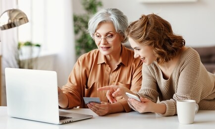 young  daughter with smartphone smiling and showing in laptop to elderly lady with credit card while shopping online at home together