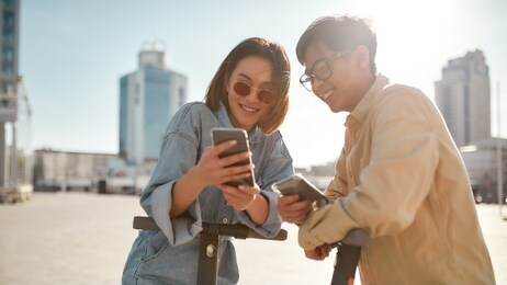 portrait of asian girl showing boy something interesting on her phone while standing outdoors. millennial guys using eco transport in downtown. modern urban lifestyle in summer. horizontal shot