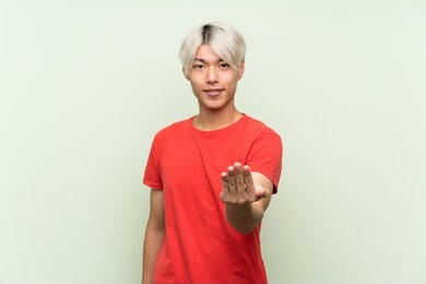 young asian man over isolated green background inviting to come with hand. happy that you came