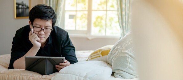 asian man lying on cozy bed using digital tablet and pen in bedroom. businessman wearing glasses working at home during quarantine. remote working or telecommuting with internet and gadget device.