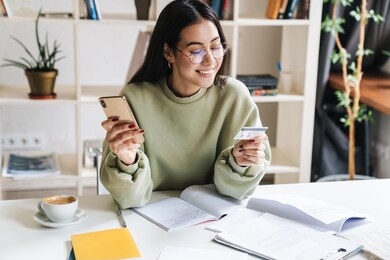 image of a beautiful happy optimistic young girl student indoors studying using mobile phone holding credit card.